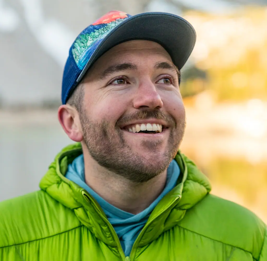 A headshot of Mac wearing a hat and a green jacket