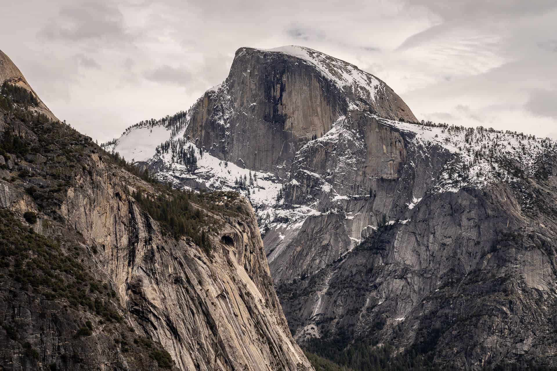 A rugged mountain peak, partially covered with snow, rises against a cloudy sky. The rocky slopes, perfect for a JMT resupply 2023 adventure, are dotted with patches of evergreen trees. The landscape appears serene and majestic.