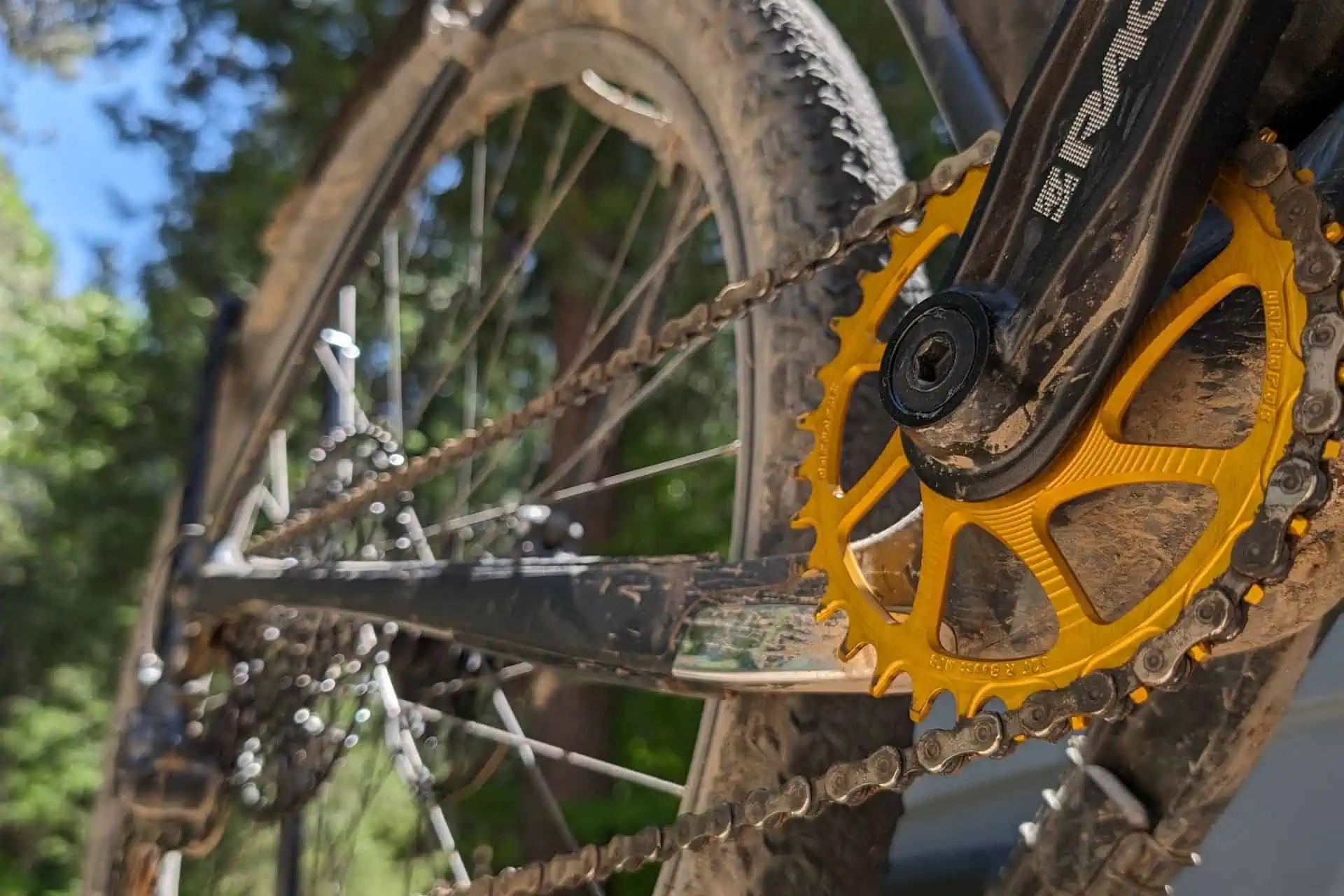 Close-up of a bicycle drivetrain with a mud-covered chainring and chain, essential for any 2024 backpacking gear list. The bike rests outdoors with blurred green foliage, hinting at an adventurous trail ride. The yellow chainring stands out against the dark, muddy components.