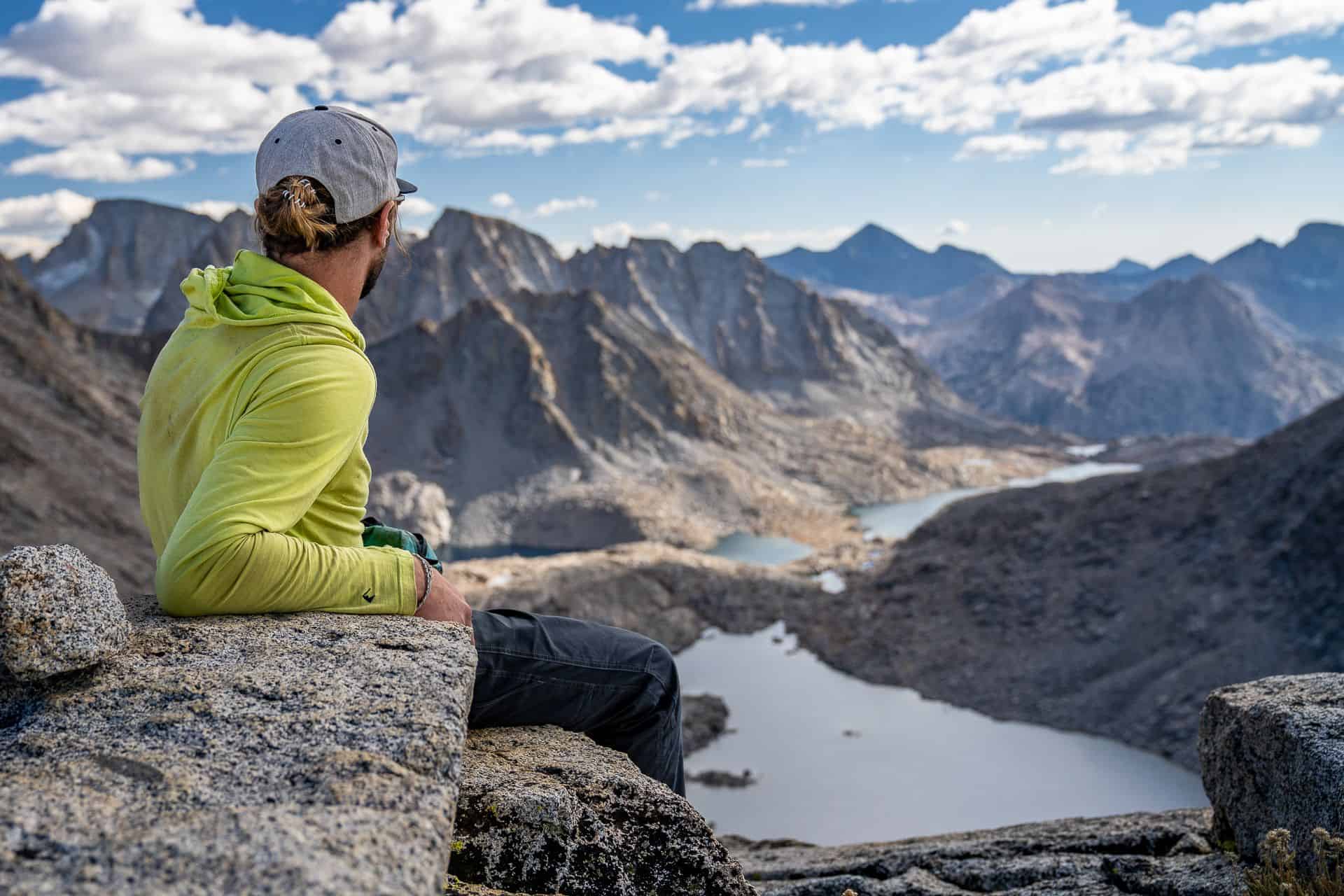 A person in a bright green jacket and gray cap sits on a rocky ledge, bridging the interesting-weird divide as they overlook a stunning mountain landscape with lakes under a partly cloudy sky. The scene is serene and expansive, suggesting tranquility and adventure.