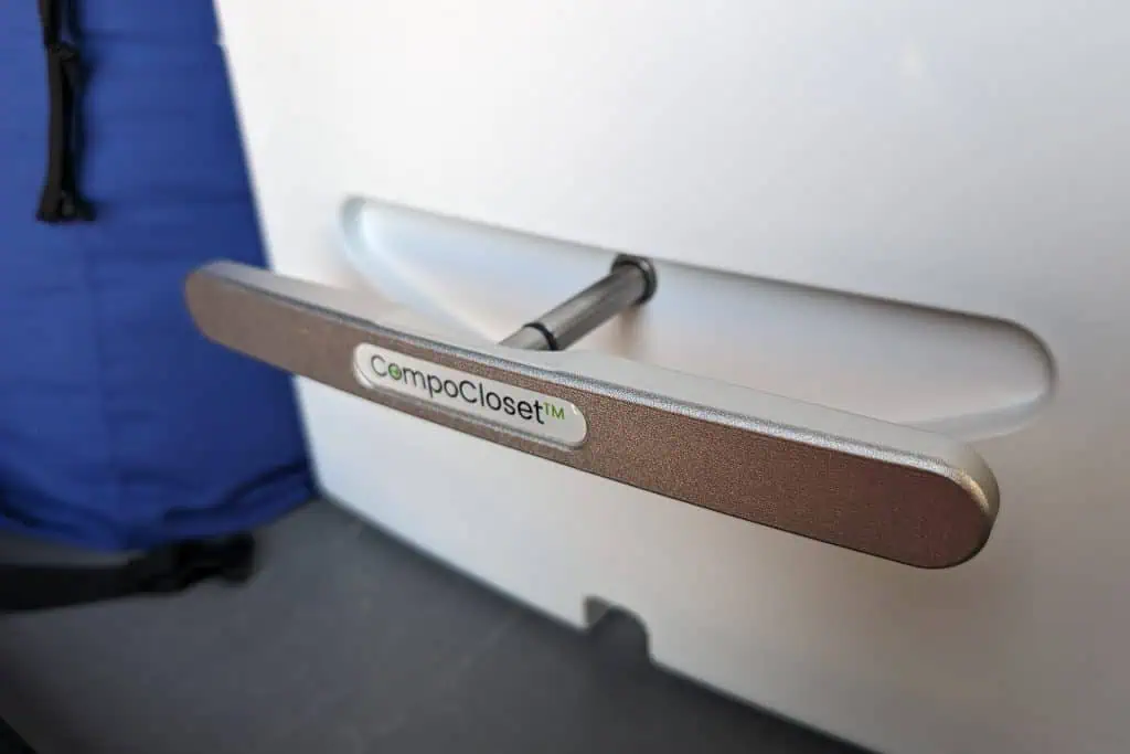 Close-up of a handle on a white CompoCloset unit, showcasing a metallic bar with a pivoting mechanism. The logo CompoCloset adorns the handle. A blue fabric item is partially visible in the background, ideal for those seeking an efficient cuddy composting toilet solution.