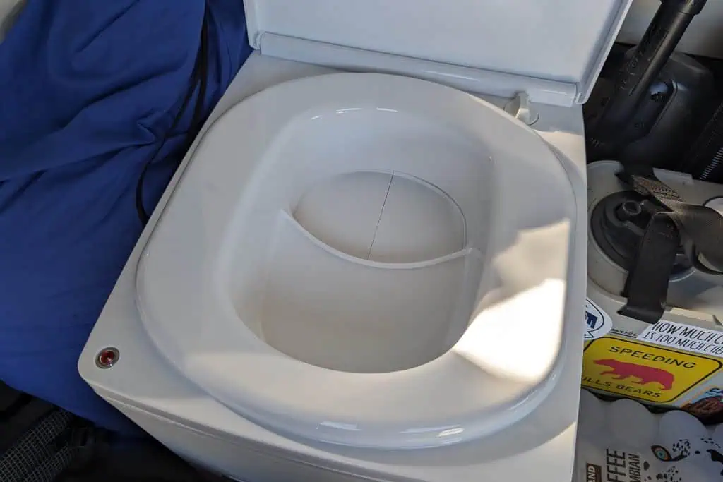 A close-up of a white cuddy composting toilet with the lid open, revealing a divided interior. A blue fabric is partially visible on the left, with assorted items, including a magazine, on the right. Sunlight casts intriguing shadows inside this innovative setup.