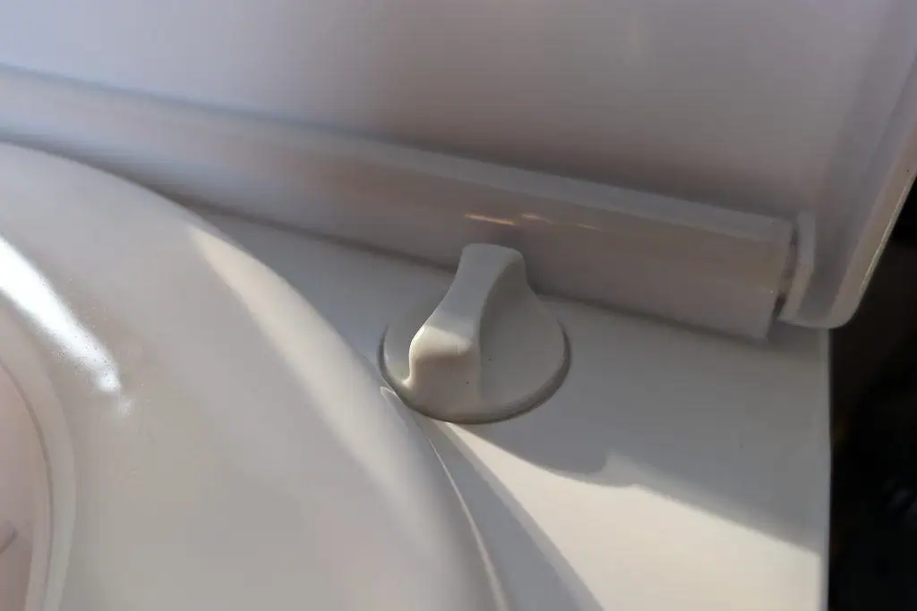 A white plastic dial, perhaps from a cuddy composting toilet, is embedded on a flat surface in this close-up. Soft shadows suggest sunlight streaming from the left, casting delicate patterns as the dial sits with a slight tilt to the right.