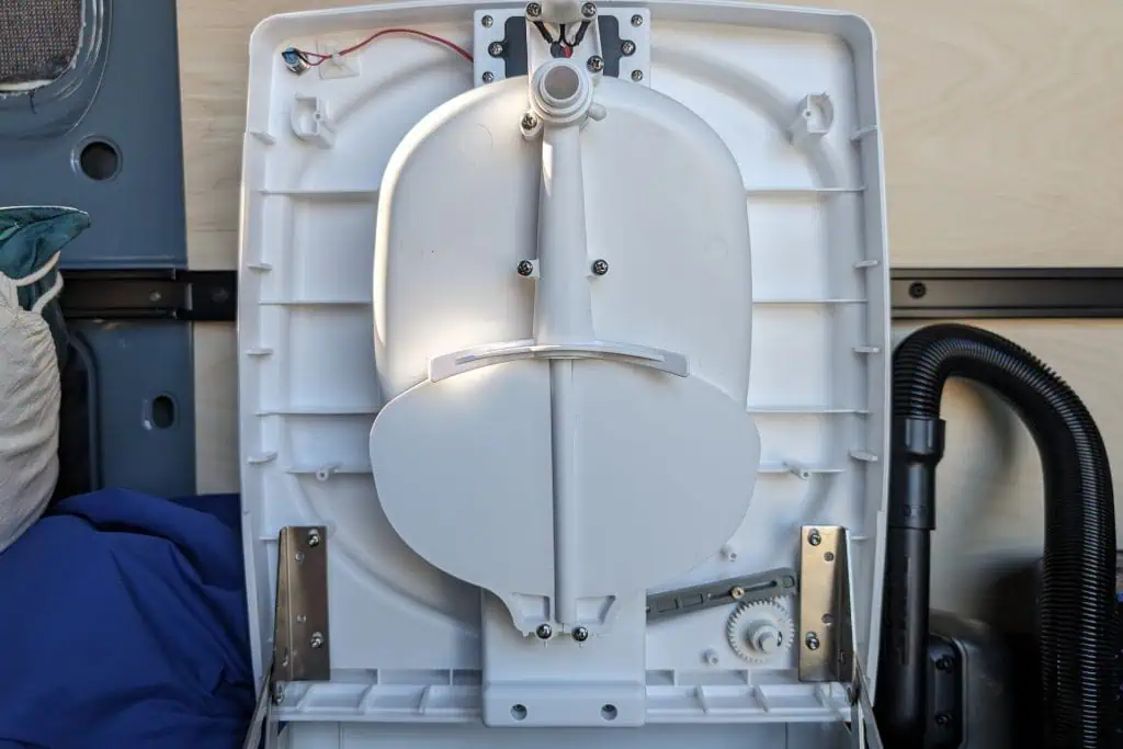 Image of an open cuddy composting toilet showing its interior components and wiring. The lid is removed, revealing mechanical parts and a circular section in the center. A hose is visible on the right, with a pillow in the background on the left, ideal for an insightful review.