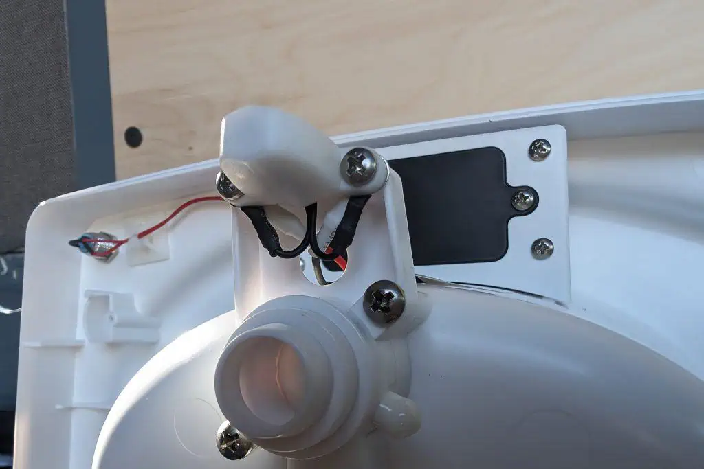 Close-up of the underside of a toilet lid, showcasing a secure bidet attachment connected with wires and a tube. The sturdy setup, reminiscent of those praised in cuddy composting toilet reviews, includes screws and a sleek black plastic component on a light-colored surface.