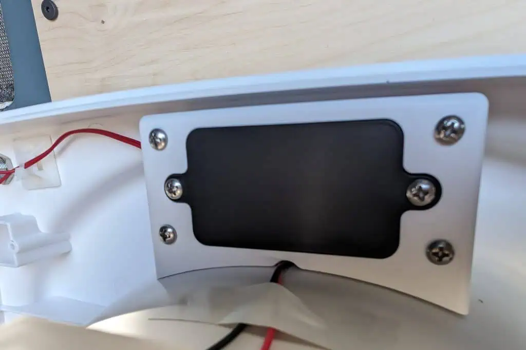 Close-up of an electronic devices interior reveals a black rectangular component secured with four screws on a white surface, reminiscent of the precise engineering seen in cuddy composting toilets. Red and black wires are connected, with part of a wooden surface visible in the background.