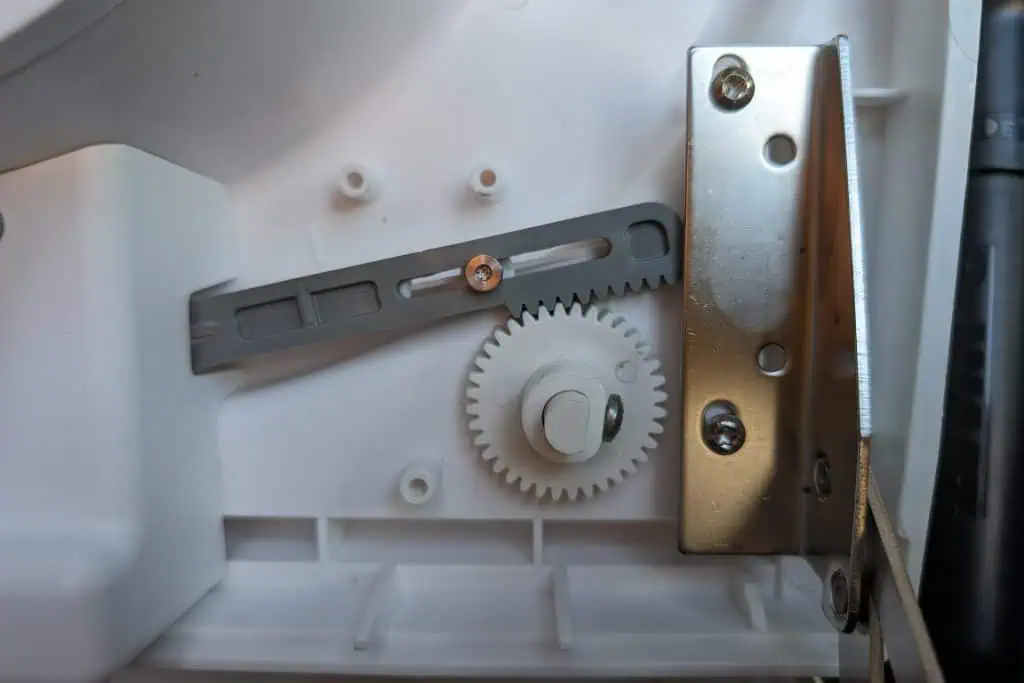 Close-up of a white mechanical component featuring a gear mechanism, reminiscent of intricate parts in a cuddy composting toilet. A gray plastic lever with a screw and metal bracket are visible on the white panel, surrounded by various screws and connection points, indicating an assembly part.