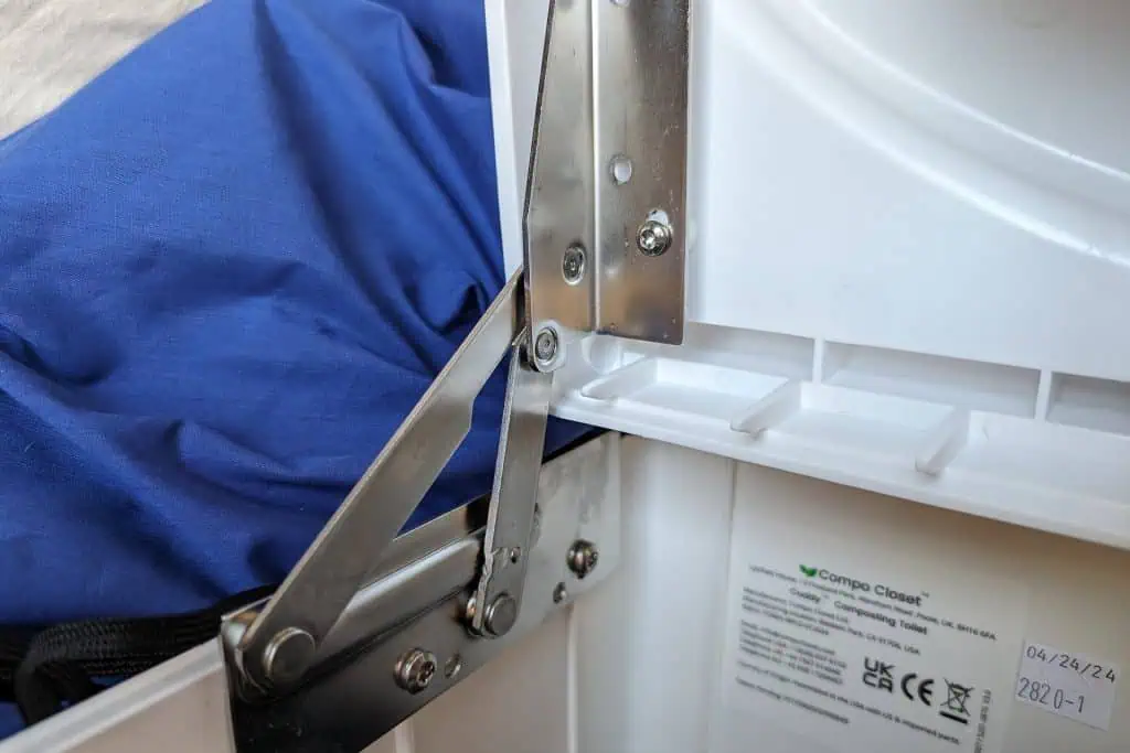 Close-up of a hinge mechanism inside a white cuddy composting toilet, with a visible label revealing manufacturing details. In the background, a folded blue fabric is partially visible, adding to the review-like assessment of this compact design.