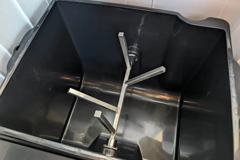 Top view of a black plastic container with a metal agitator inside, resembling a cuddy composting toilet. The agitator, featuring angled blades on a central rod, is surrounded by white tiles, creating reflections in the lighting.