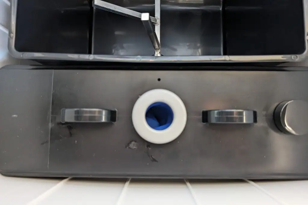 Close-up of a black rectangular object with two handles, a circular opening in the middle lined with blue, and a knob on the right. This cuddy composting toilet features a metallic component above it, possibly part of a larger machine or appliance.