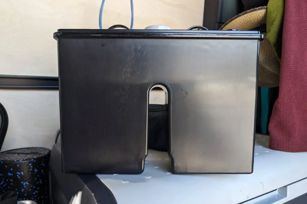 A black plastic rectangular container with an opening at the bottom sits on a flat surface. Wires are visible above it, resembling components of a cuddy composting toilet. Fabric items surround the area, including a patterned cylindrical object and a red curtain.