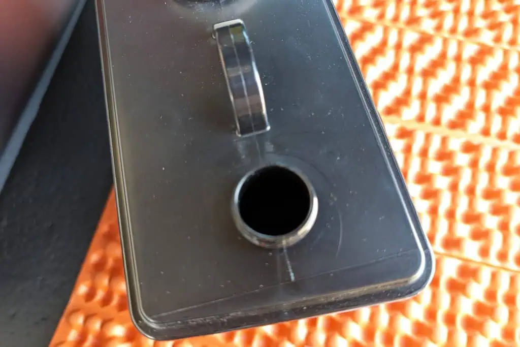 Close-up of a black plastic container with a spout, positioned on an orange textured surface. Resembling a component from a cuddy composting toilet, the open spout faces the viewer, inviting inspection.