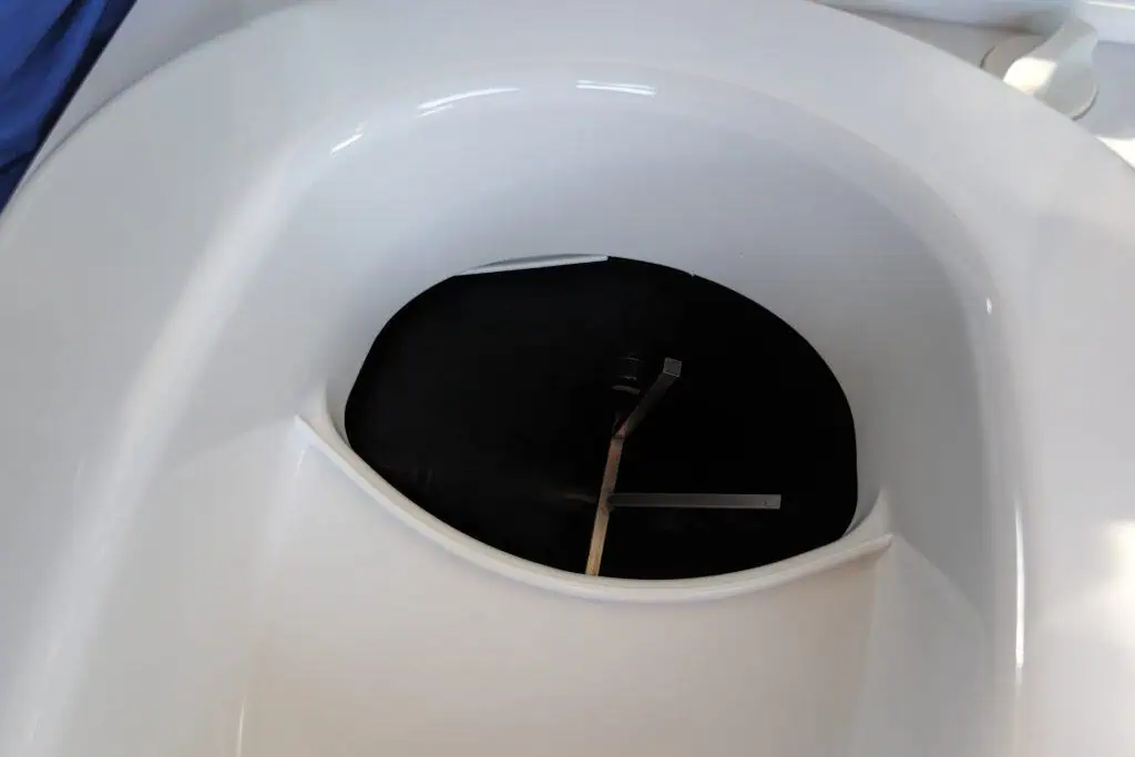 A view of an airplane toilet with the lid raised reveals the dark, circular opening of the waste compartment, reminiscent of a compact cuddy. The smooth white surroundings and a hint of blue fabric in the corner complete the efficient design.