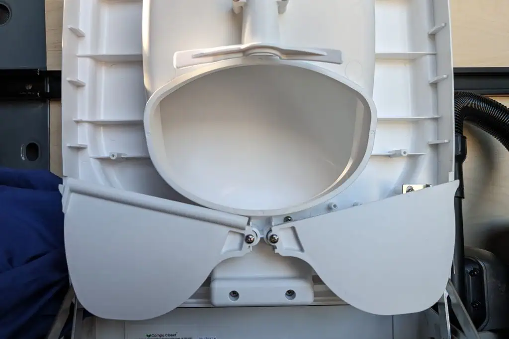 Close-up of an open white toilet lid and seat in an upright position, revealing the inner bowl. The surrounding area includes plumbing components and a glimpse of blue fabric on the left, reminiscent of a scene youd find in a cuddy composting toilet review.