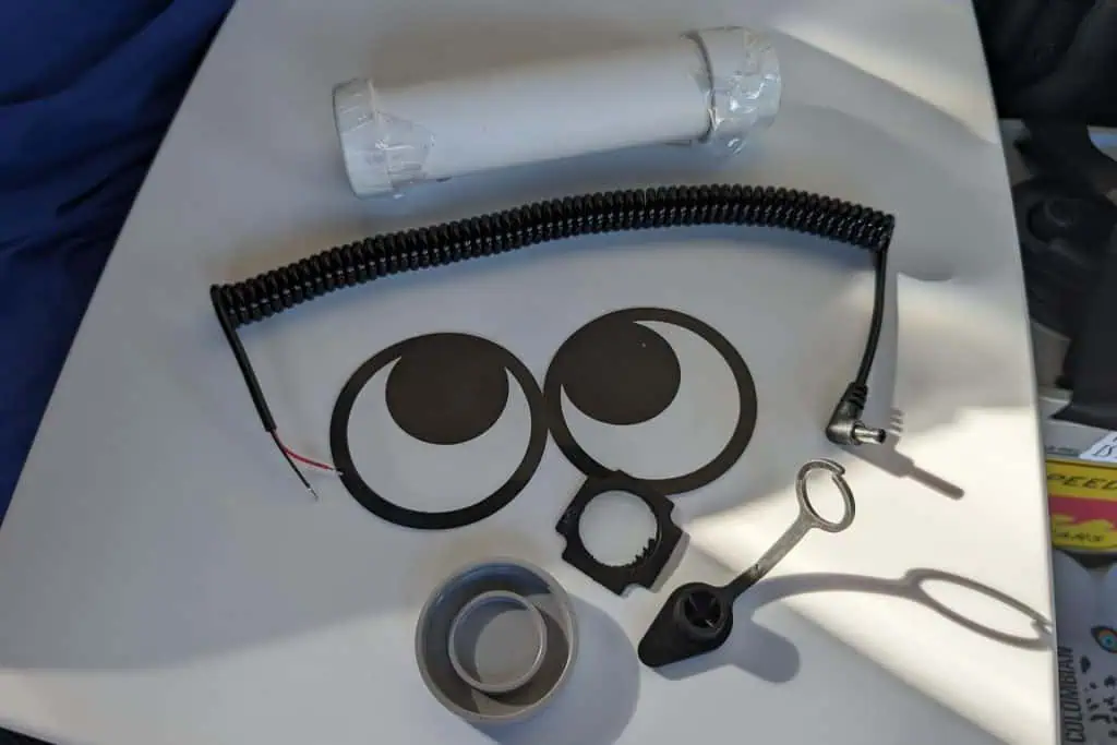An assortment of electronic components on a white surface, reminiscent of a DIY kit you might find in a detailed cuddy composting toilet review, includes a coiled cable, round metallic rings, mechanical parts, and a small cylindrical object wrapped in white paper. Shadows from the components are visible.