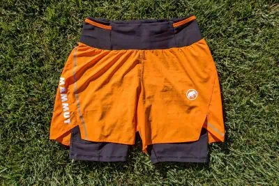 Orange and black Mammut Aenergy TR 2 in 1 athletic shorts with a dual-layer design, featuring the brand logo and name on the left side. The shorts are laid flat on green grass, highlighting a reflective strip elegantly visible on the outer layer.