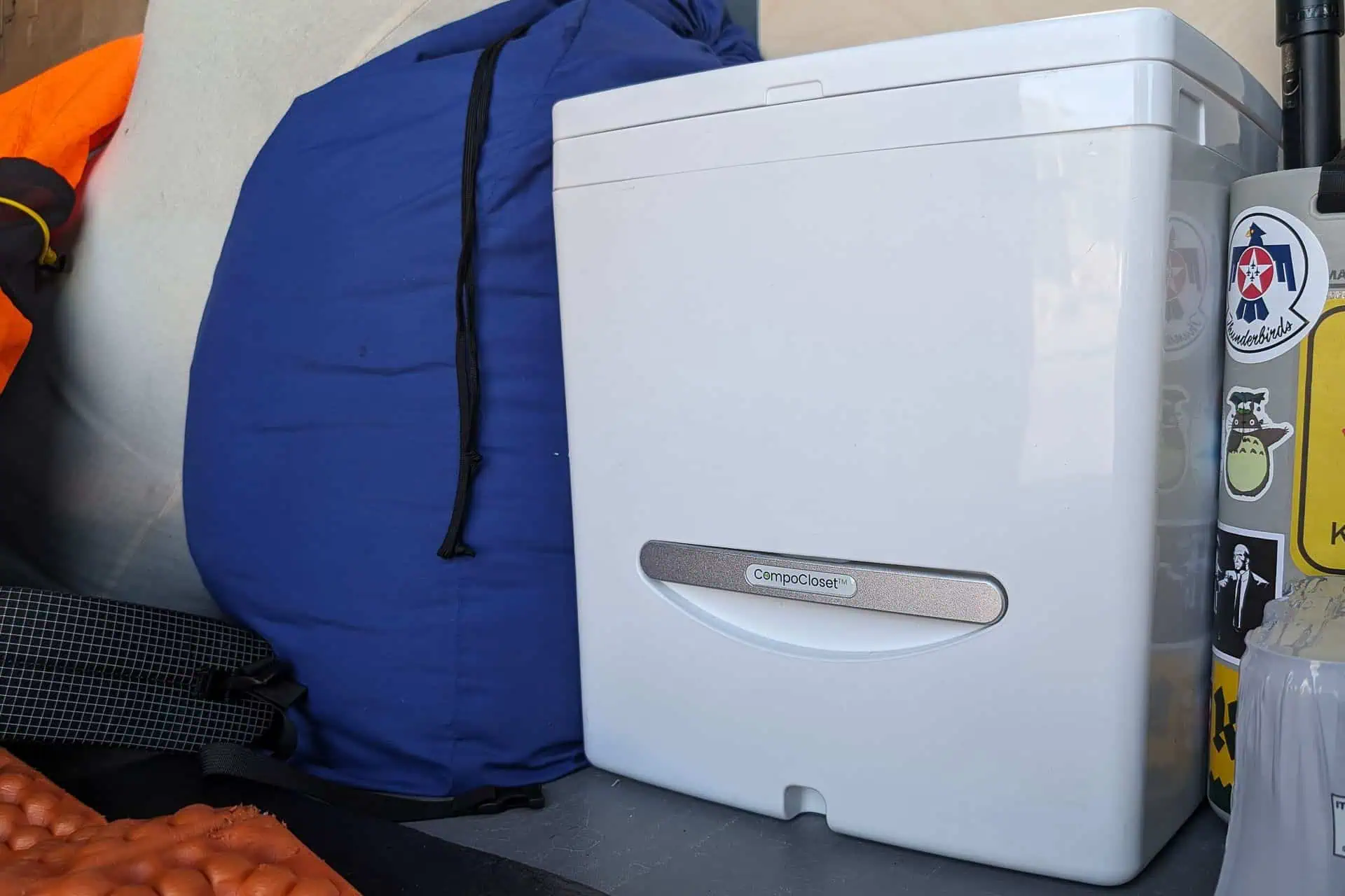 A portable washing machine sits beside a large blue duffel bag and an orange sleeping mat, reminiscent of a detailed cuddy composting toilet review. The machine is white with a silver handle, surrounded by various items like a black mesh bag and stickers on nearby surfaces.
