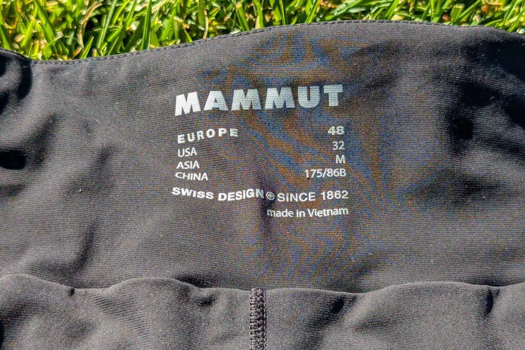 Black Mammut clothing label on fabric, featuring sizes: Europe 48, USA 32, Asia M, China 175/88A. Text reads: Swiss Design Since 1862, and Made in Vietnam. Set against green grass, these are the Mammut Aenergy TR 2 in 1 Shorts.