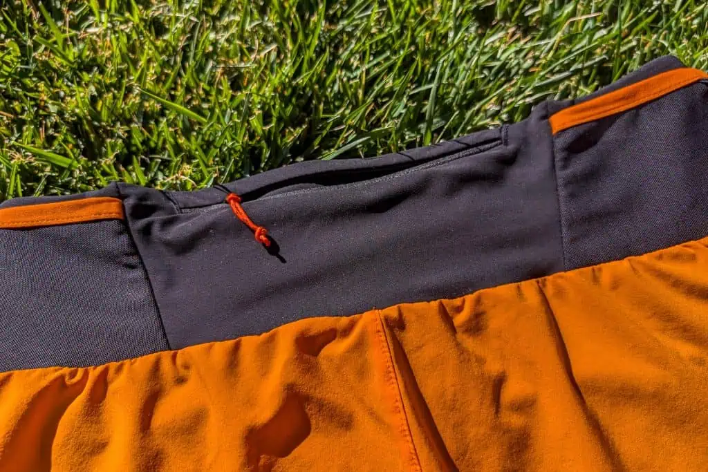 Close-up of an orange and black fabric item, possibly Mammut Aenergy TR 2 in 1 Shorts, lying on green grass. A small red loop is visible at the black fabrics edge. The texture of the shorts is highlighted by the sunlight.