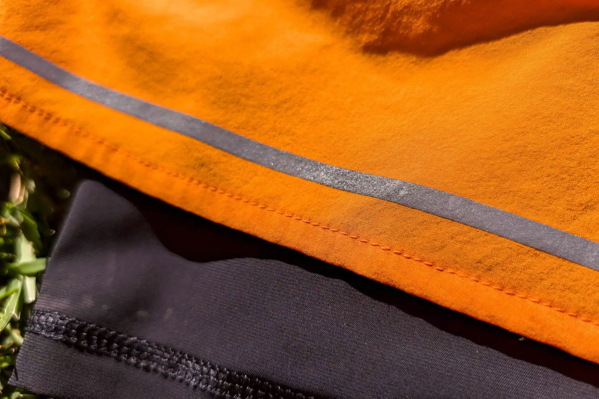 A close-up showcases the texture and contrast of the Mammut Aenergy TR 2 in 1 Shorts, featuring an orange fabric with a reflective silver stripe overlapping black fabric with visible stitching. A hint of grass peeks in, adding a touch of nature to this dynamic scene.