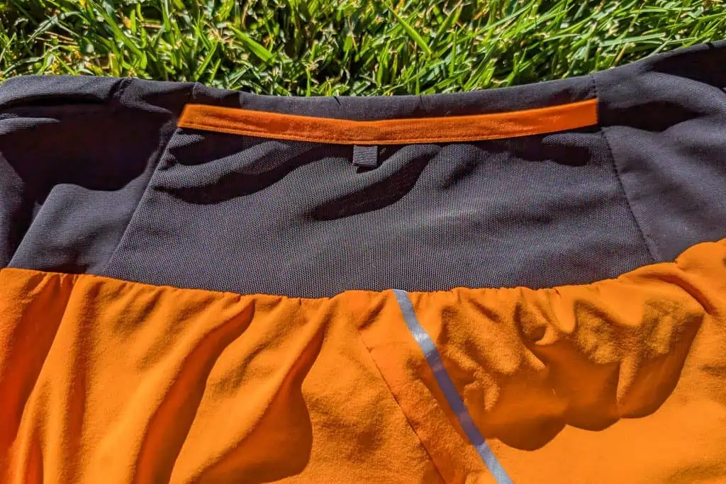 Close-up of an orange and black Mammut Aenergy TR 2 in 1 Shorts spread on green grass. The garment features a ruched design with a reflective strip on the orange section, contrasting the vibrant and dark hues beautifully.