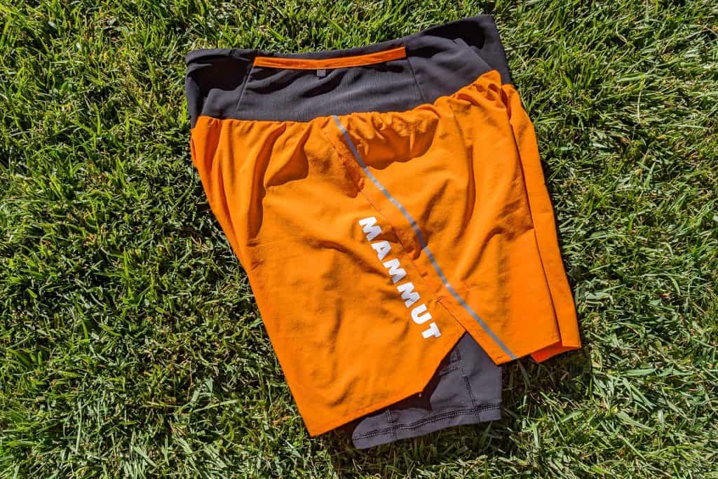 Orange and black Mammut Aenergy TR 2 in 1 Shorts with a blue stripe, neatly folded on green grass.