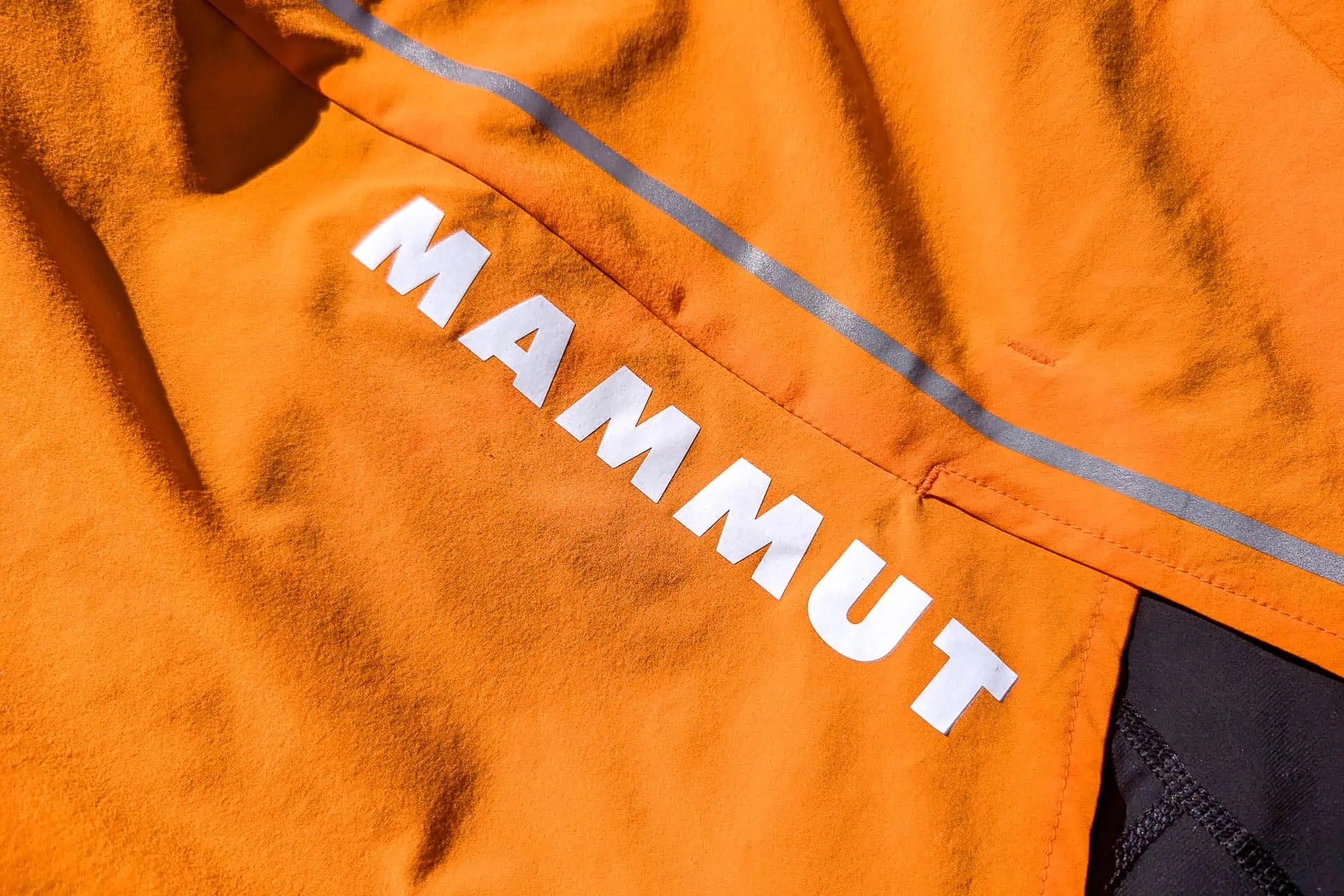 Close-up of an orange fabric, likely from Mammut Aenergy TR 2 in 1 Shorts, with MAMMUT in bold white letters. A reflective gray stripe runs diagonally across the textured surface, suggesting its part of durable outdoor gear.