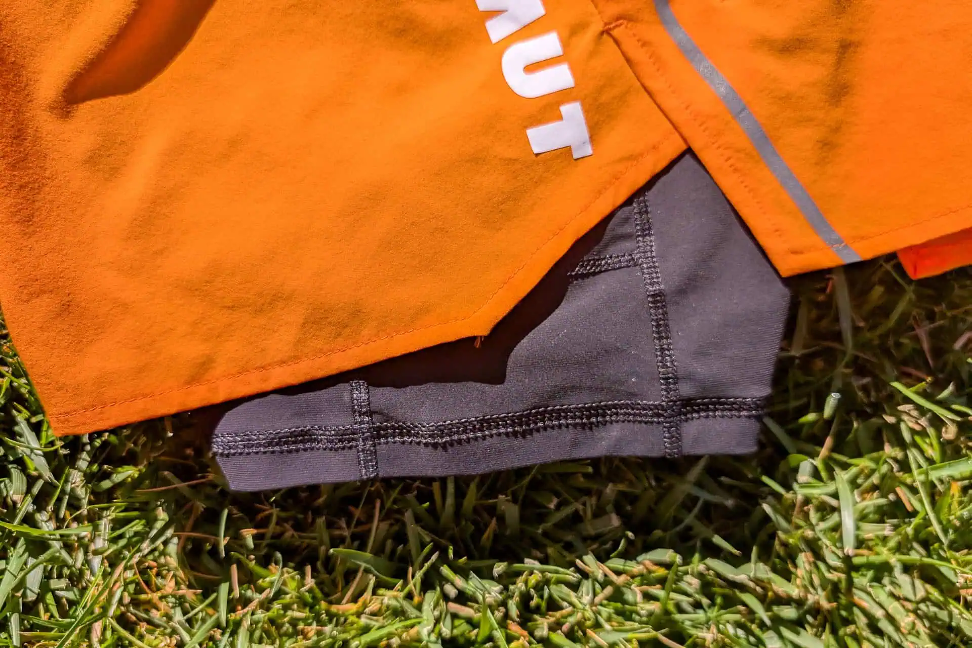 Close-up of orange Mammut Aenergy TR 2 in 1 Shorts featuring a white partial logo overlaid on black compression shorts. Lying on lush green grass, the visible stitching and reflective strip on the vibrant fabric highlight their athletic appeal.