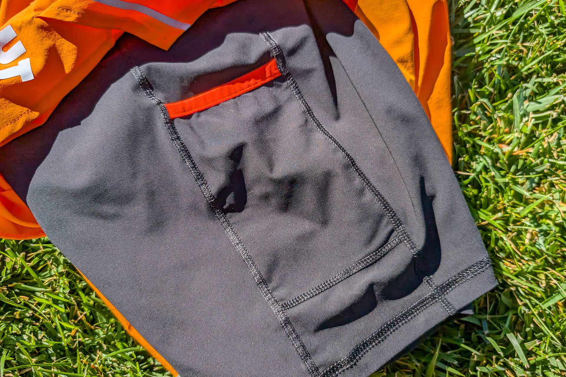 A close-up of a black cycling jersey pocket with red trim and visible stitching lies on the green grass, beside a vibrant orange section. Its contrasting colors mirror the dynamic design seen in Mammut Aenergy TR 2 in 1 Shorts, bringing style and functionality together seamlessly.