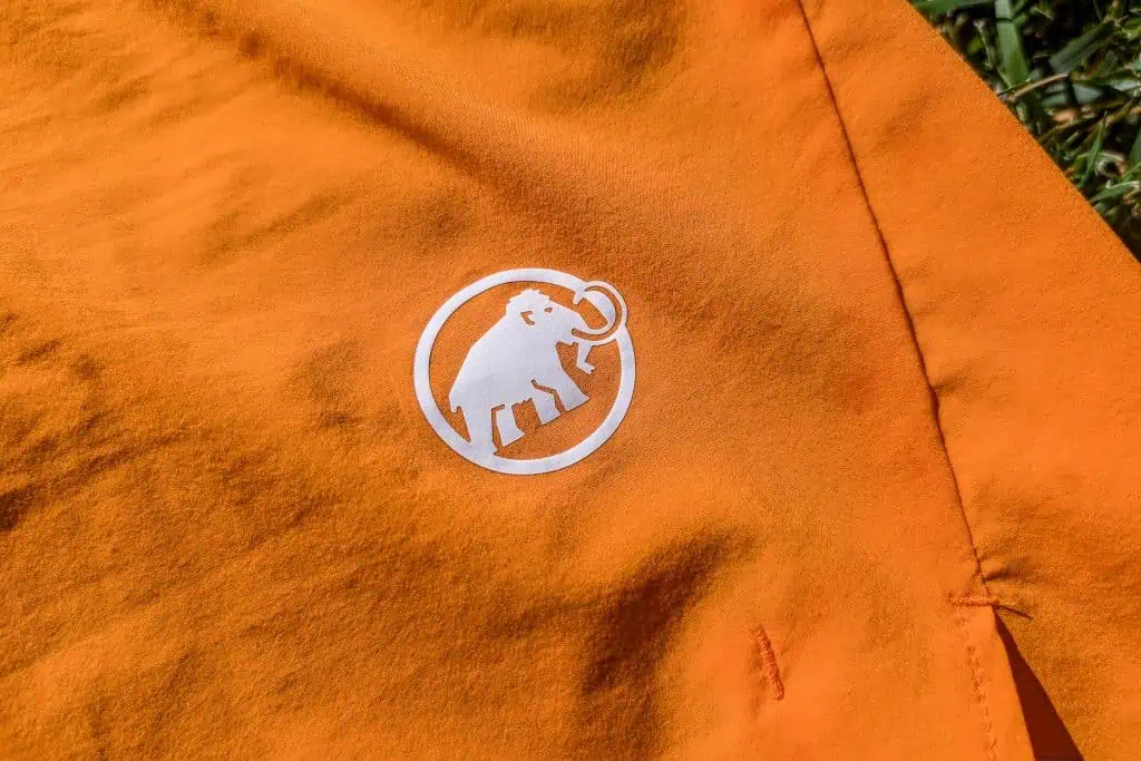 Close-up of an orange fabric with a white logo featuring a mammoth inside a circle. The fabric, likely from Mammut Aenergy TR 2 in 1 Shorts, appears to be part of outdoor clothing, placed on a grassy background.