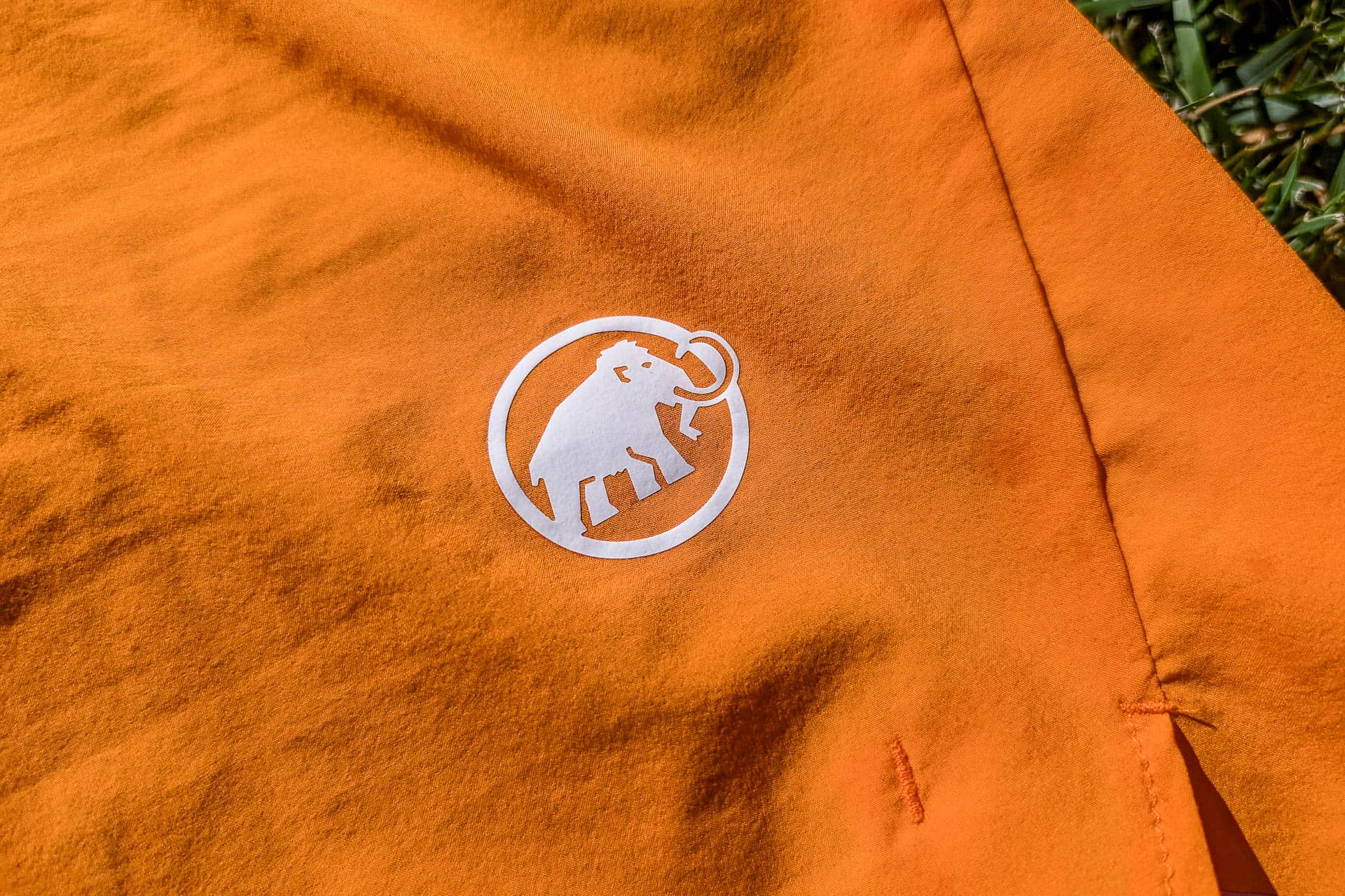 Close-up of an orange fabric with a white logo featuring a mammoth inside a circle. The fabric, likely from Mammut Aenergy TR 2 in 1 Shorts, appears to be part of outdoor clothing, placed on a grassy background.