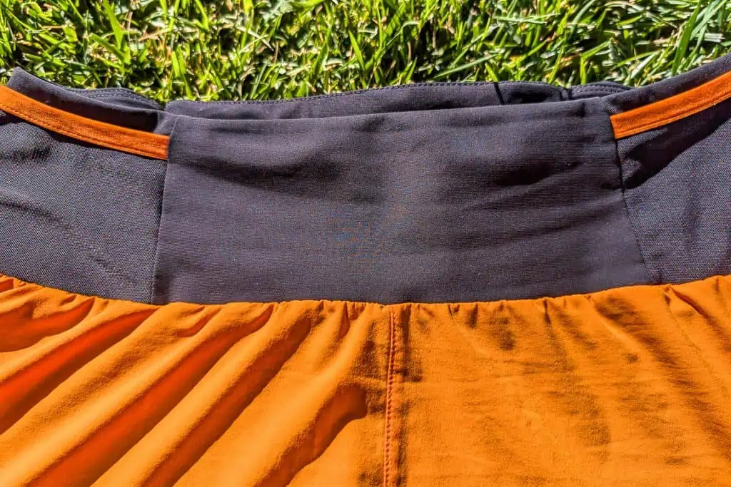 Close-up of an orange and black Mammut Aenergy TR 2 in 1 Shorts lying on grass, highlighting the waistband area. The fabric is slightly gathered at the orange section, creating soft folds and texture. The grass is bright green, indicating a sunny day.
