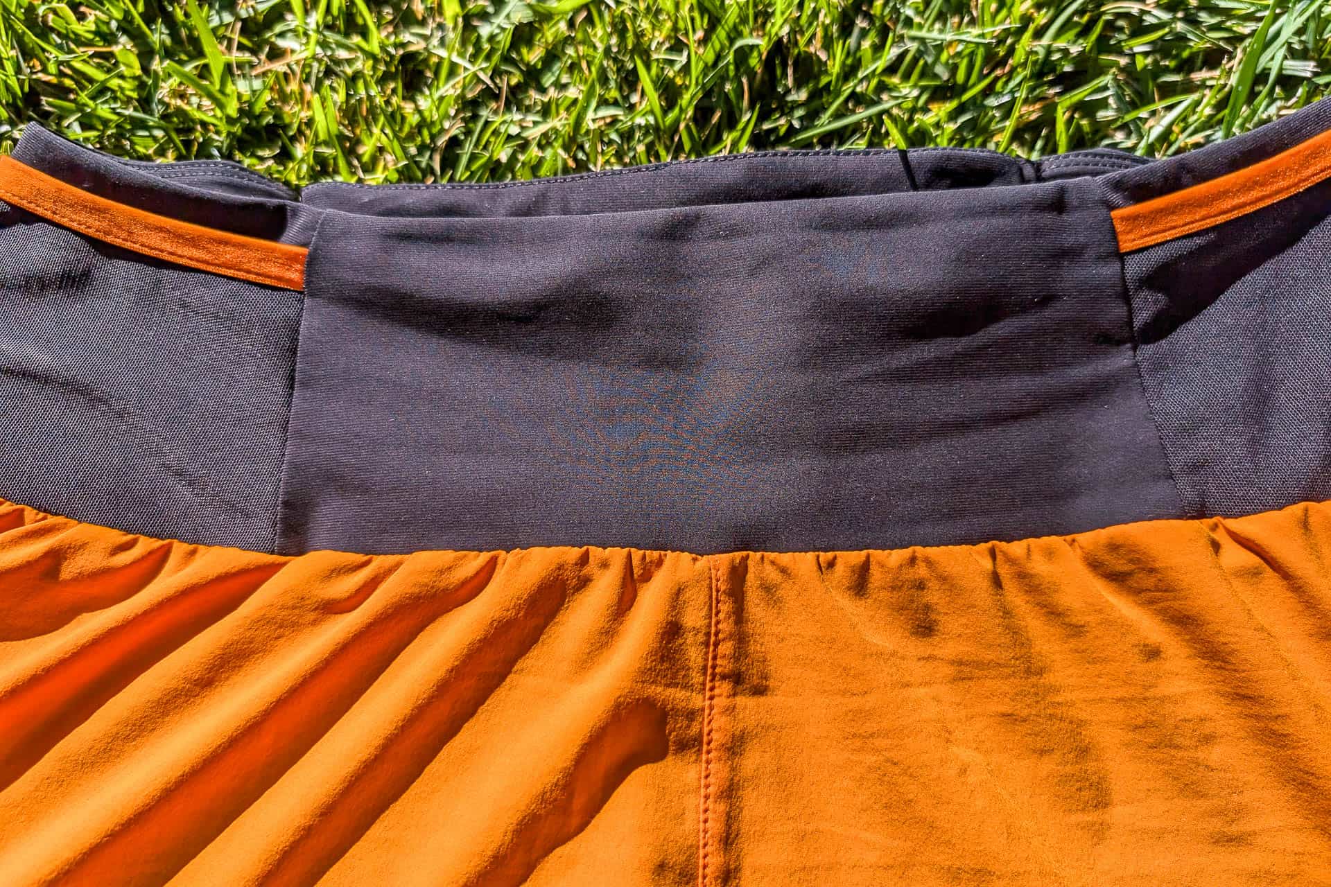 Close-up of an orange and black Mammut Aenergy TR 2 in 1 Shorts lying on grass, highlighting the waistband area. The fabric is slightly gathered at the orange section, creating soft folds and texture. The grass is bright green, indicating a sunny day.