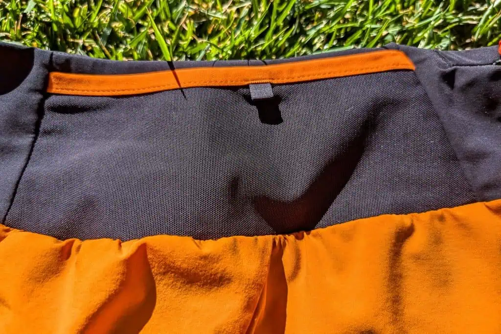 Close-up of Mammut Aenergy TR 2 in 1 Shorts with a black waistband and orange fabric, lying on green grass. Sunlight highlights the texture and colors, creating a contrast between the dark and bright hues.