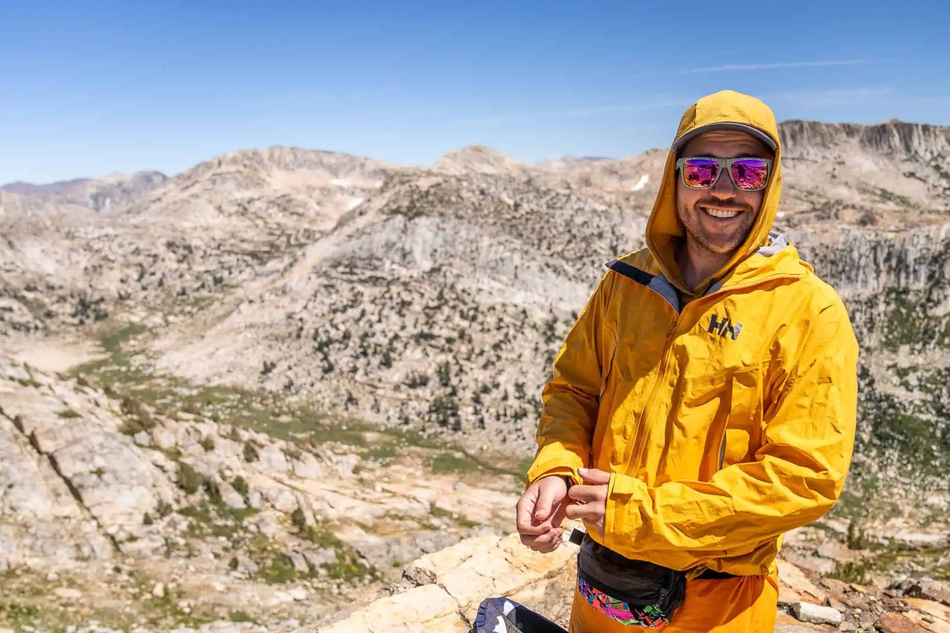 A person wearing a Helly Hansen Verglas Infinity Shell Jacket 2.0 and sunglasses smiles while standing on a rocky mountain landscape. The background features expansive, rugged terrain with clear blue skies.