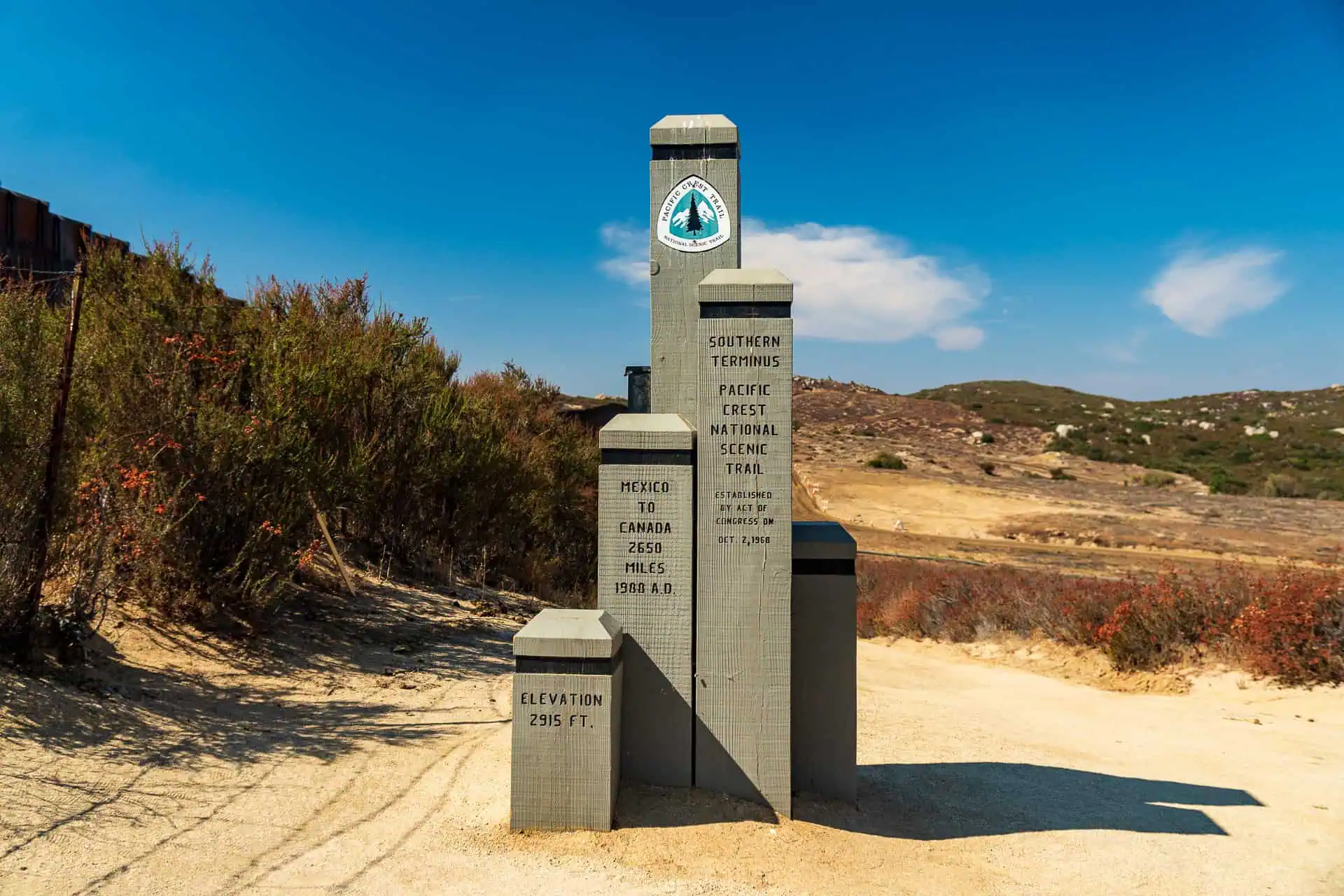 A stone monument marks the southern terminus of the Pacific Crest National Scenic Trail, with inscriptions detailing the trails mileage and elevation. Surrounded by dry vegetation and hills under a clear blue sky, it stands ready for those embarking on upcoming pct surveys in 2024.