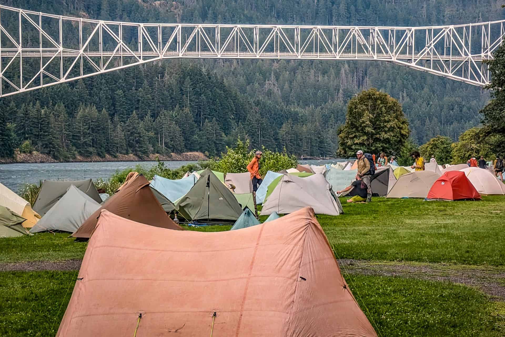 A campsite with multiple tents is set up on grassy ground, perfect for relaxing after exploring the best thru-hiker gifts. In the background, a large bridge spans a river, with dense forested hills rising beyond. Campers are visible near the tents.