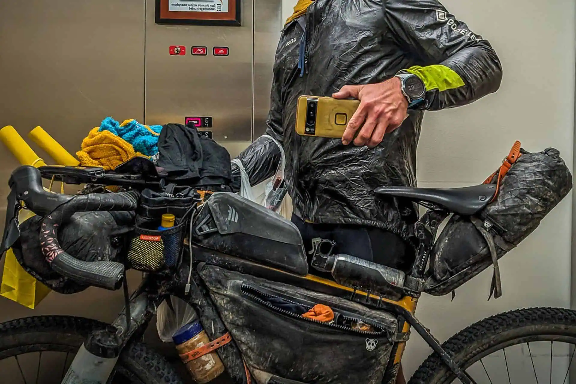 A person in a rain jacket takes a mirror selfie inside an elevator, holding their smartphone. Accompanied by their favorite gear for the Tour Divide, they stand beside a heavily packed bicycle with multiple bags, a water bottle, and various items strapped to it.