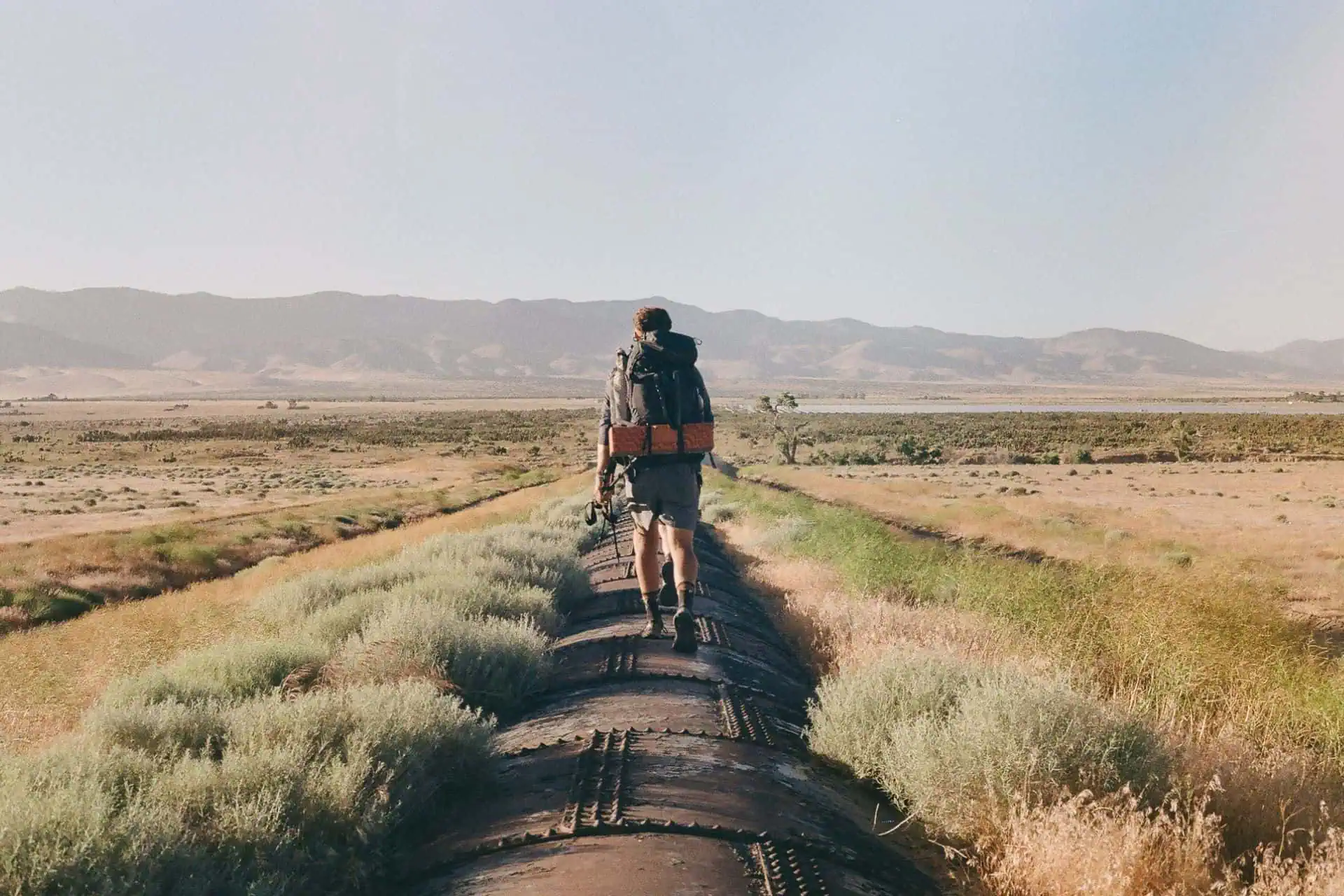 A person with a backpack, armed with the latest PCT gear guide 2025, navigates a narrow trail or pipeline through a dry, grassy landscape. Mountains rise in the distance beneath a clear sky.