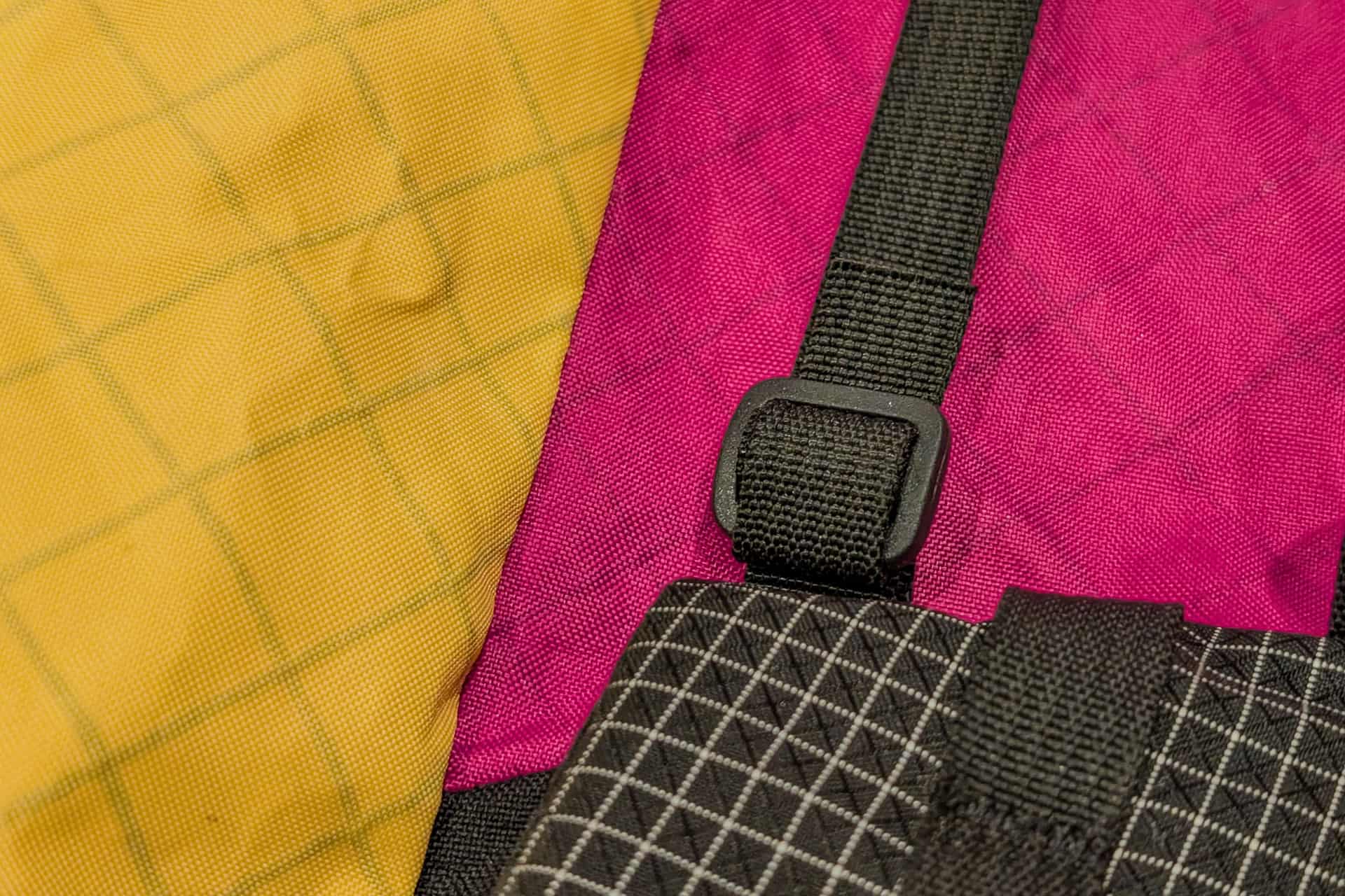 Close-up of colorful Atom Packs Atom backpack fabric, featuring a yellow checkered pattern on the left, a magenta section in the middle, and a black and white grid pattern on the right. A black strap with an adjuster crosses over the fabric.
