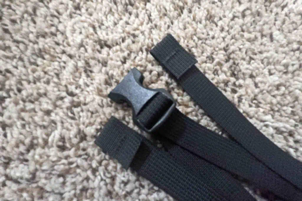 Three black nylon straps with a black plastic buckle lie on a beige textured surface, likely carpet. The buckle, reminiscent of the design seen in Atom Packs Prospector gear, consists of two parts attached to each strap, ensuring a secure clip together.