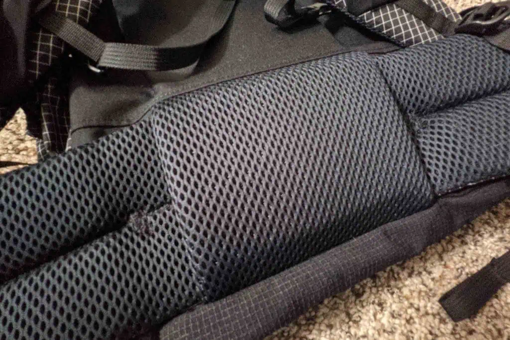 Close-up of an Atom Packs Prospector backpacks padded and textured back panel with black mesh material. The rectangular padded sections ensure comfort, while the straps peek from the top against a brown, textured backdrop.