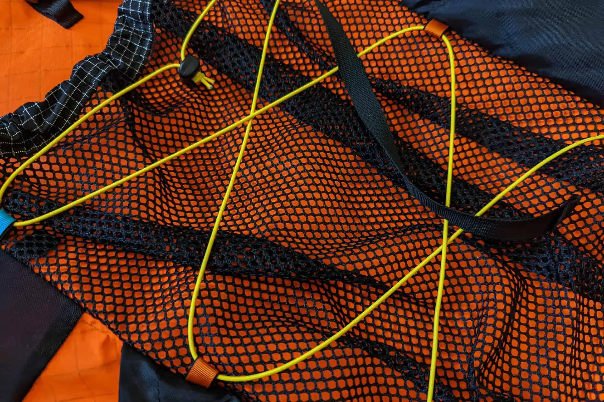 Close-up of an Atom Packs Pulse item featuring orange and black mesh fabric with yellow cords crisscrossing over it. The netted pattern, a hallmark of outdoor gear like backpacks, is complemented by adjustable toggles on the cords.