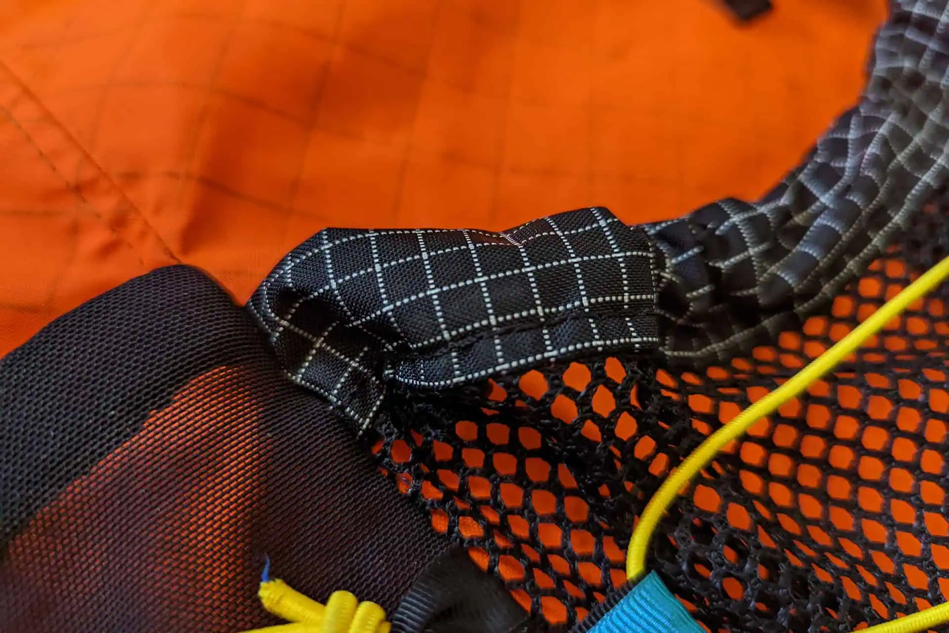 Close-up of an orange and black fabric from the Atom Packs Pulse series, featuring a black checkered strap, black mesh, and yellow and blue cords. The materials are layered and interwoven, showcasing fine stitching and detailed texture.