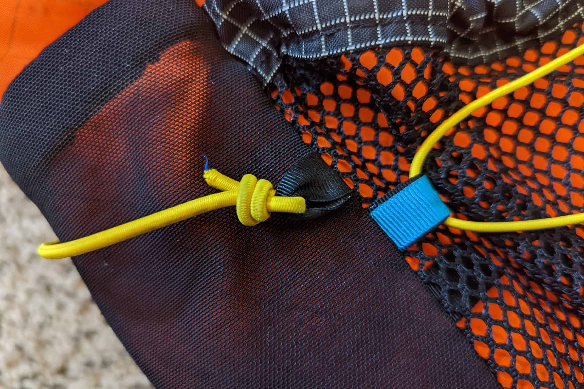 Close-up of a black mesh fabric with an orange lining, reminiscent of the sleek Atom Packs Pulse design. A yellow cord is tied in a knot, and a blue tag is attached to it. The fabric features a distinctive grid pattern at the top.