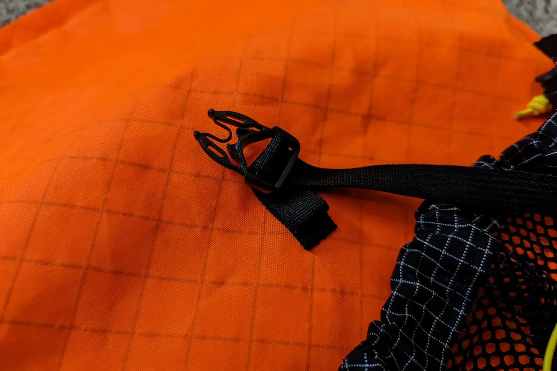 Close-up of a black buckle strap on an orange Atom Packs Pulse fabric with a grid pattern, attached to a black and white mesh material. The background is a soft surface, possibly cloth or carpet.