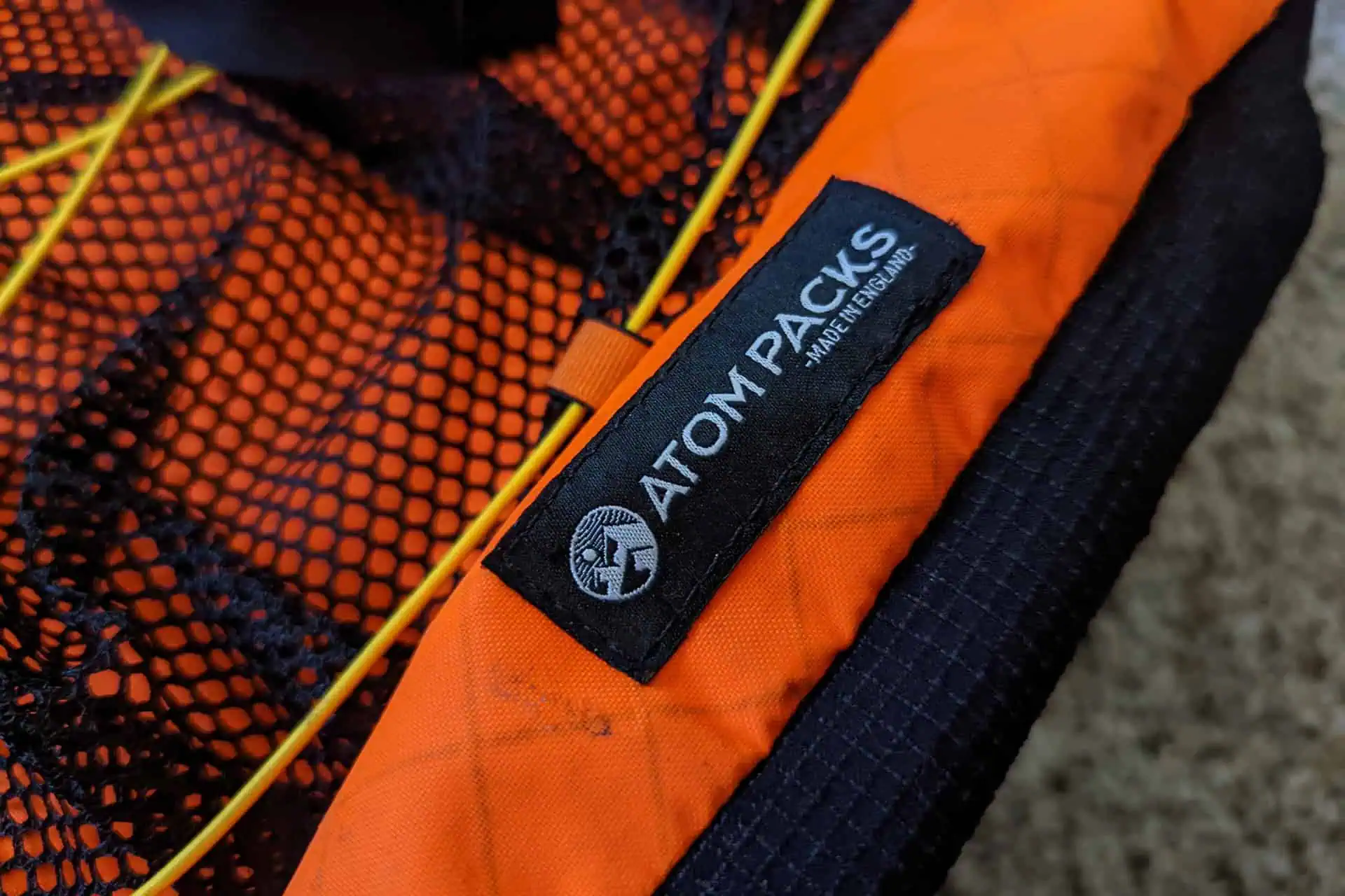Close-up of a backpack showcasing an orange fabric and black mesh design. The black label proudly reads ATOM PACKS Pulse, complete with a circular logo. Yellow bungee cords add contrast, set against a textured grey surface.