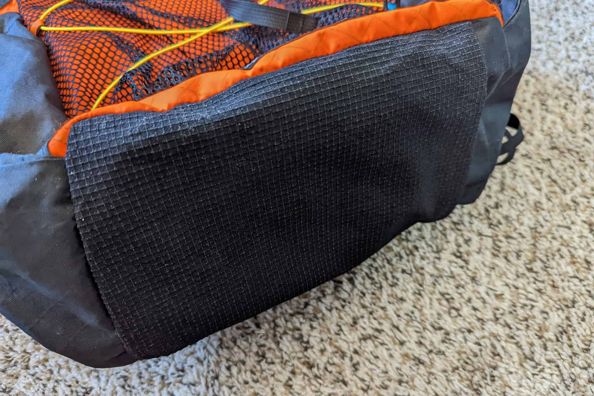 Close-up of an Atom Packs Pulse backpack corner resting on a carpet. The pack showcases an orange and black color scheme with mesh and textured fabric, while yellow cords add distinction to the mesh section.