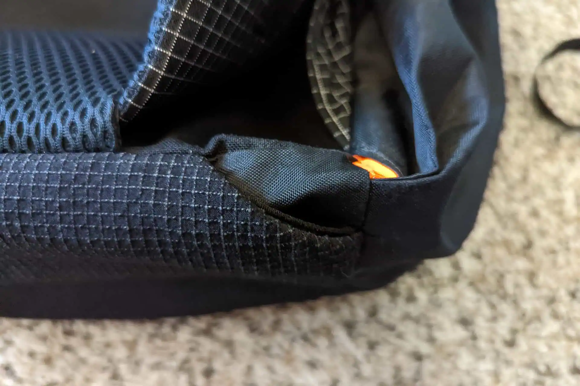 Close-up of a black Atom Packs Pulse backpack corner, showcasing textured fabric and mesh material with an orange loop or tab. The bag rests on a light-colored carpet.