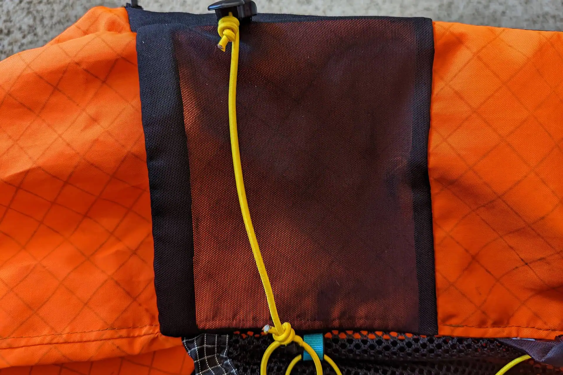 Close-up of an Atom Packs Pulses orange fabric with a diamond pattern, featuring a black rectangular patch. A yellow cord with a stopper is threaded through the patch. Below, a section of black mesh material is visible.